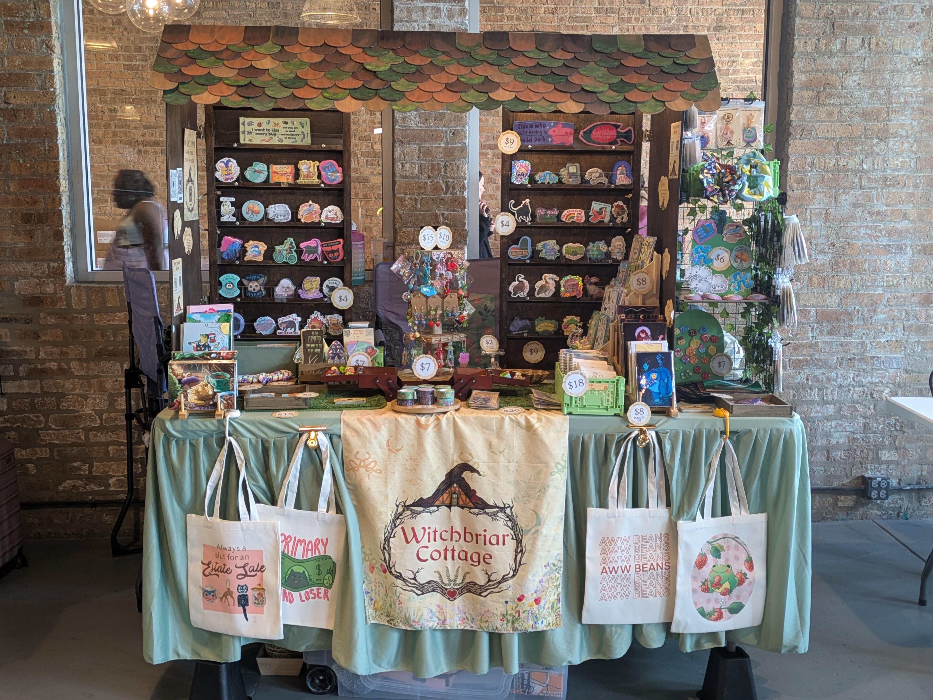 Witchbriar Cottage booth at a market, with a shingled roof, shelves of stickers and pins, and tote bags hanging from a draped green table.
