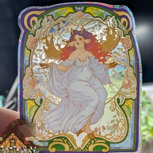 Holographic suncatcher featuring an Art Nouveau illustration of a deer girl with antlers and flowing red hair in a white dress, framed by ornate stained-glass-style floral borders in purple and gold