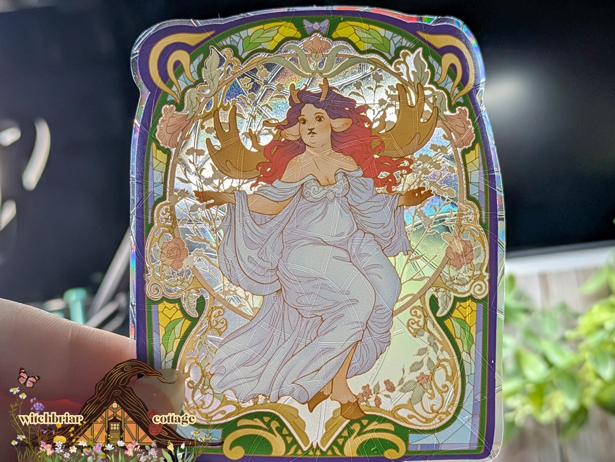 Holographic suncatcher featuring an Art Nouveau illustration of a deer girl with antlers and flowing red hair in a white dress, framed by ornate stained-glass-style floral borders in purple and gold