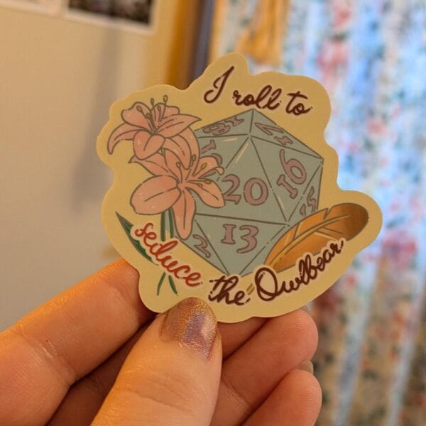 Die-cut sticker of a D20 die decorated with flowers and feathers, with the text 'I roll to seduce the Owlbear'